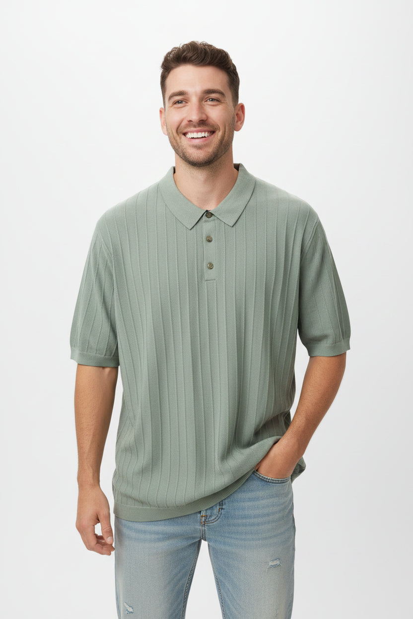 A smiling man wearing a sage green ribbed short-sleeve polo shirt and light blue jeans, standing against a plain light gray studio background.