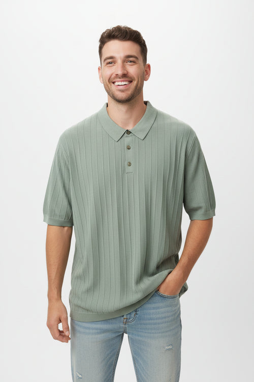 A smiling man wearing a sage green ribbed short-sleeve polo shirt and light blue jeans, standing against a plain light gray studio background.