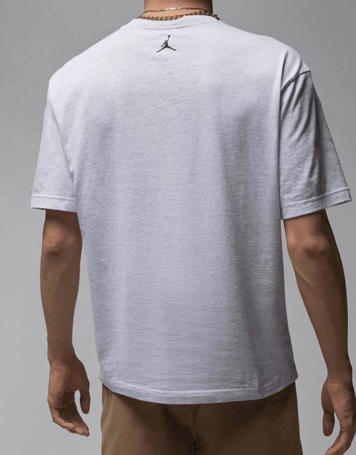 A man wearing a light gray heathered t-shirt from the back, featuring a small black Jumpman logo centered below the collar. The model is also wearing brown pants and a beaded necklace, set against a solid gray studio background.