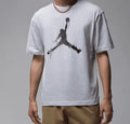 A man wearing a light gray t-shirt with a black distressed Jumpman graphic on the front, paired with khaki pants and a gold chain necklace, against a neutral gray background.