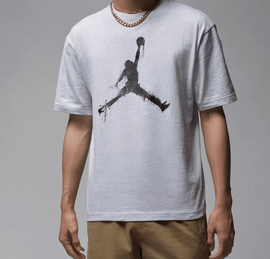 A man wearing a light gray t-shirt with a black distressed Jumpman graphic on the front, paired with khaki pants and a gold chain necklace, against a neutral gray background.