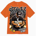 An orange short-sleeve graphic t-shirt featuring a large front print of a man in a black cap reading 'GOAT' and holding stacks of cash, with stylized text reading 'STACKS' and 'PAID IN FULL'.