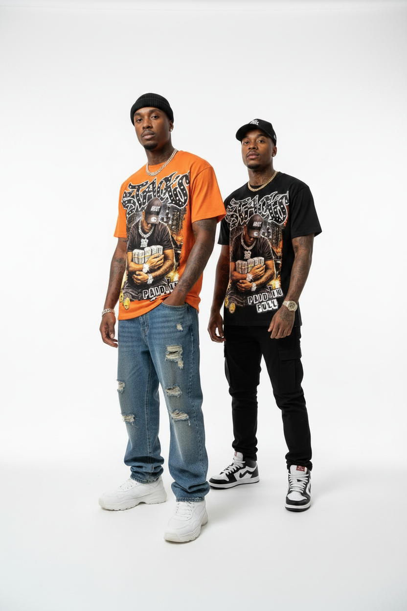 Two men modeling graphic t-shirts against a plain white studio background. The man on the left wears an orange t-shirt with a large graphic print and distressed blue jeans, while the man on the right wears a black t-shirt with a similar graphic print and black pants.